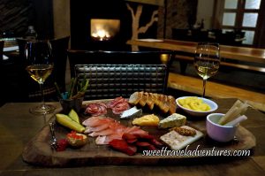 A Wooden Board Sitting on a Wooden Table With a Variety of Meats, Cheeses, Pickled Vegetables and Beans, and Slices of Bread With A Glass of White Wine on Each Side of the Board, and a Lit Fireplace in the Background