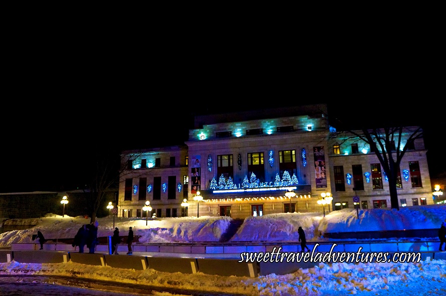 An Outdoor Ice Rink at Night With a Few Ice Skaters, Snow Piled Up Around the Rink, Behind it a Tree on the Right and One on the Left and Further Behind a Large Building With Decorative Lights Lit Up