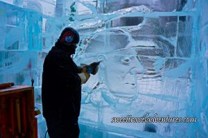 A Man With a Black Winter Coat and a Black Touque Using a Tool to Shape the Ears of a Man Wearing a Baseball Cap Whose Face and Head is Etched Into an Ice Wall, a Wooden Box With Tools Hanging Inside it to the Left of the Ice Sculptor
