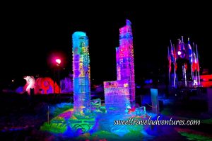 Ice Sculpture of Skyscrapers Lit Up at Night With Multiple Coloured Lights, in the Background on the Right is Several Poles With Flags and on the Left is a Snow Sculpture Lit Up