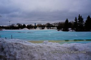 A Frozen Blue Pond With Snow Piled in Front and in the Background Two Teepees and Trees and a Cloudy Grey Sky