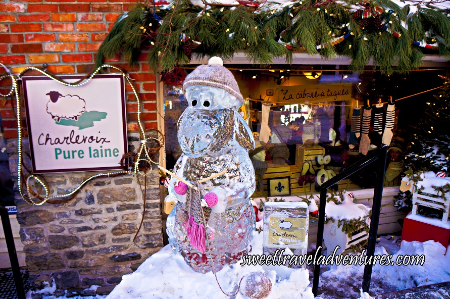 A Lamb Ice Sculpture Wearing a Tan Touque and Holding Knitting Needles and a Half Knit Tan and Pink Scarf, Standing in Front of a Brick Building With a Large Glass Window Looking Into a Store With Items on Display and Green Garland Hanging Above the Window, and a Tan Coloured Square Store Sign to the Left of the Window Hanging from the Brick Building Which Has Orange Brick on the Upper Half and Light Brown Brick on the Lower Half