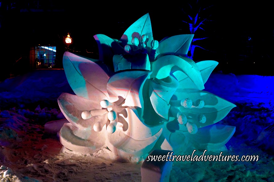 A Snow Sculpture of a Group of Three Maple Flowers at Night Lit Up With a White Light, a Green Light, and a Little Bit of Purple Light, Behind the Sculpture is Dark Blue Lights on the Snow. and a Building in the Background on the Left