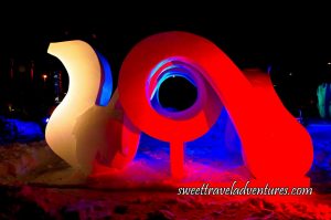 A Snow Sculpture at Night That Looks Like a Vertical Squiggle With a White Light on it Attached to a Circular Structure With a Piece That Leads Downwards and is Lit Up With a Red Light, and Snow Underneath the Sculpture and Behind it