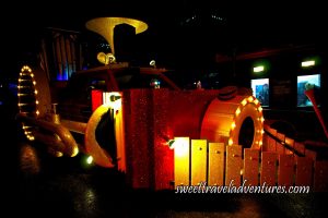A Red Truck With Red Sparkles on the Street at Night Made to Look Like a Train, on the Front is a Yellow Drum With Lights Around it and Around the Drum Are Two Yellow Xylophones Forming a Triangular Shape on the Front of the Train, a Yellow Horn Sticking Out the Top, a Yellow Trombone Along the Side of the Train With Lights Lit Up Around the Circular Part of the Instrument, a Yellow Organ Sticking Out the Back, and a Structure on the Other Side of the Street With Lights on and People Inside the Two Windows on the Right