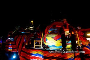 A Float at Night with Musicians Sitting on Wooden Chairs Playing Music Dressed With Red and Black Checkered Jackets and Black Pants, a Giant Ball on One End of the Float With Different Colours of Fabric Wrapped Around it, and the Arrowhead Sash Wrapped Around the Bottom of the Float