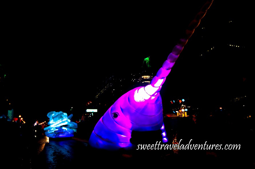 A Float on the Street at Night Shaped Like the Head of a Narwhal and Lit Up With Purple Lights and a Little Bit of Dark Blue Light, Following Behind is an Iceberg Float Lit Up With Turquoise and White Lights, in the Background are Buildings With Only a Few Lights on