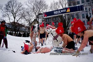 People in Their Bathing Suits Wearing Giant Red Rooster Hats Playing in the Snow With Bonhomme and Some People Lying and Rolling Around in the Snow