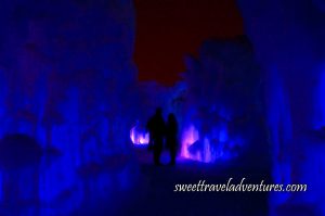 A Silhouette of a Couple Standing on a Snowy Path With Blue Frozen Ice Structures Lining the Path and in Front of Them, and the Ice Structure in Front of Them and to the Right Partially Illuminated Pink and the Night Sky Above