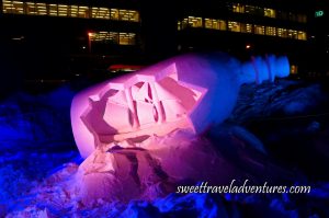 A Snow Sculpture of a Bottle Tilting Upwards to the Right on the Snow With the Front of the Bottle Cut Out and a Large Ship Showing Inside With Pink Light on the Bottle and Some Dark Blue Light, and Around it Dark Blue Snow, in the Background a Large Building With the Lights on Inside