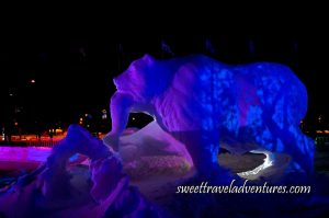 A Snow Sculpture of a Large Bear Holding a Fish in its Mouth and Some Pink Light Reflected onto the Upper Portion of the Bear and a Design of Dark Blue and Blue Lights Reflected onto the Bear's Back, Snow All Around, and Flags on Flag Poles Waving in the Wind in the Background