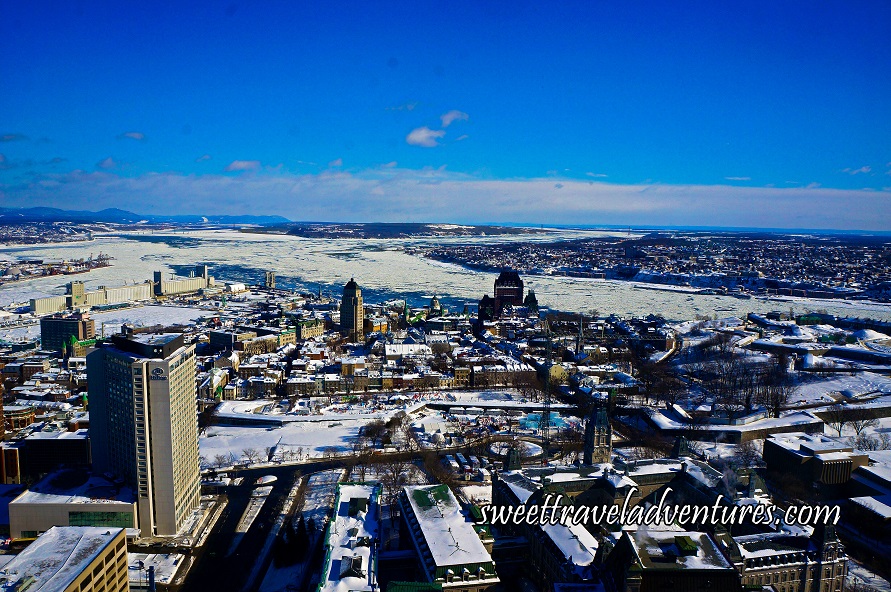 A Tall Brown Building on the Left and Other Buildings Around it, Many of Which Have Roofs Covered in Snow and the Ground is Covered in Snow, in the Distance is a River Going From the Left to Right and is Mostly Covered in Snow, Behind the River is More Buildings in the Distance and to the Left in the Distance is Mountains, there is a Blue Sky With a Lot of White Clouds on the Horizon