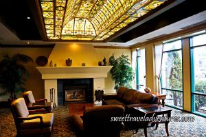 A Fireplace With a Wooden Coffee Table in Front and a Brown Sofa to the Right and Chairs on the Other Sides, Large Glass Windows on the Right, a Stain Glass Roof, and a Patterned Carpet