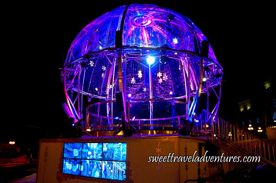 A Platform With a Giant Sphere at Night Lit Up With Purple Lights and Sparkly Stars Hanging Down From the Top of the Sphere, a Screen on the Front Side of the Platform, and Stairs Leading to the Platform on the Right