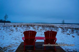 Two Red Chairs on a Wooden Platform Looking Out to a Snowy Field With a Single Bare Tree on the Right and Left and Trees in the Background and a Light Blue Sky