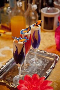 Three Champagne Glasses on a Silver Tray Filled With a White Foamy Cocktail With a Purple or Red Colour Shaped Like a Flower Around it and Topped With a Butterfly