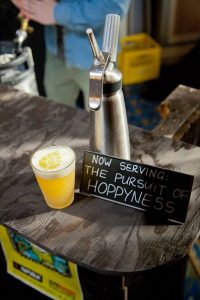 A Glass Sitting on a Grey Bar With a Bright Yellow Cocktail and One Inch of a White Foam on the Top With A Hop Drawn in the Foam