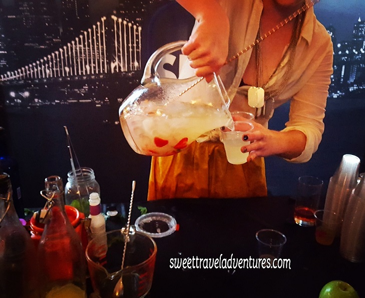 A Woman Pouring a Lemonade Cocktail With Strawberries into a Plastic Cup From a Glass Pitcher Over a Dark Table With Mixology Things, in the Background is Pictures of Cityscapes and a Bridge Lit Up at Night